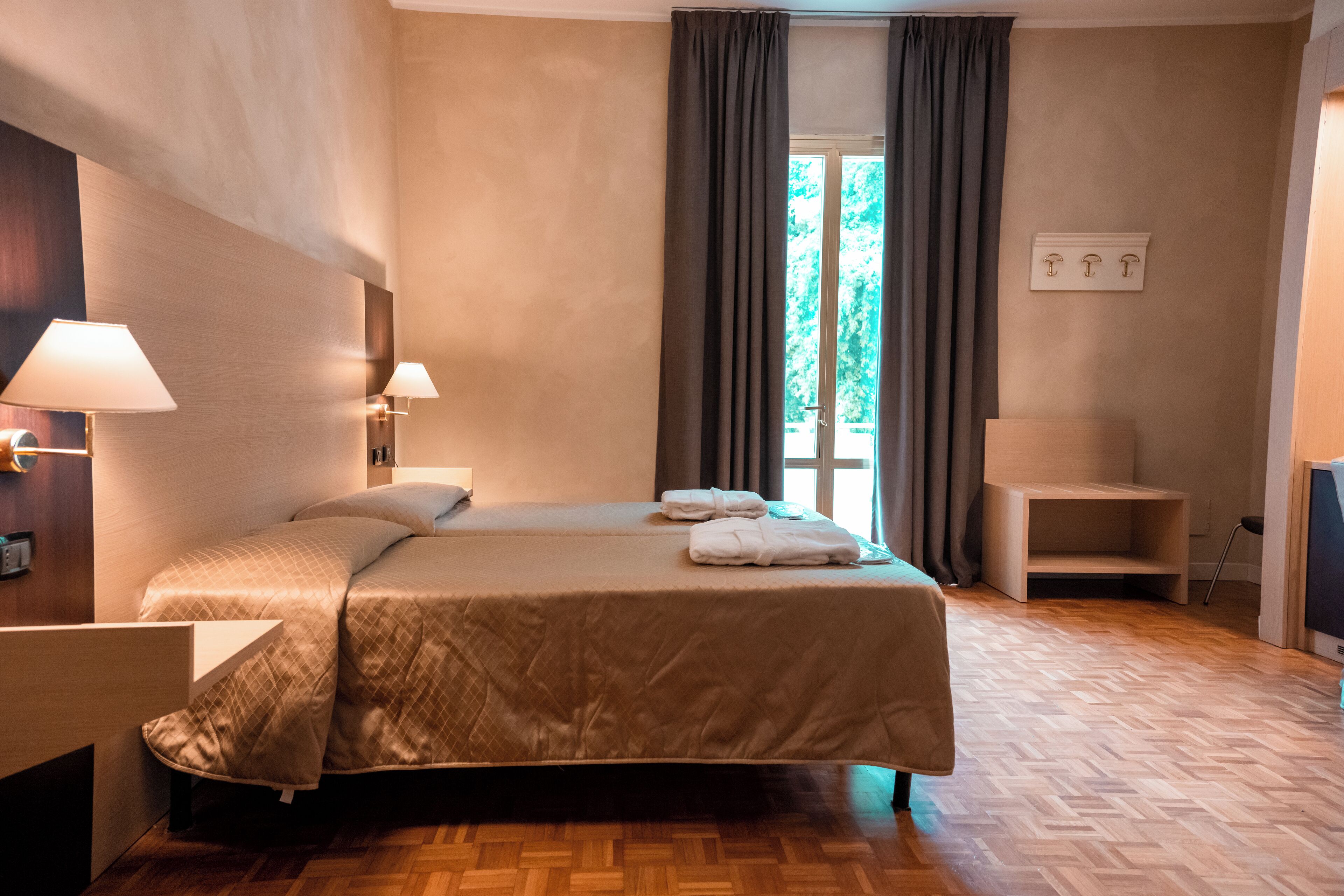 Habitación de hotel con una cama grande, dos lámparas de noche, piso de madera, una mesita y vistas a zonas verdes por la ventana.