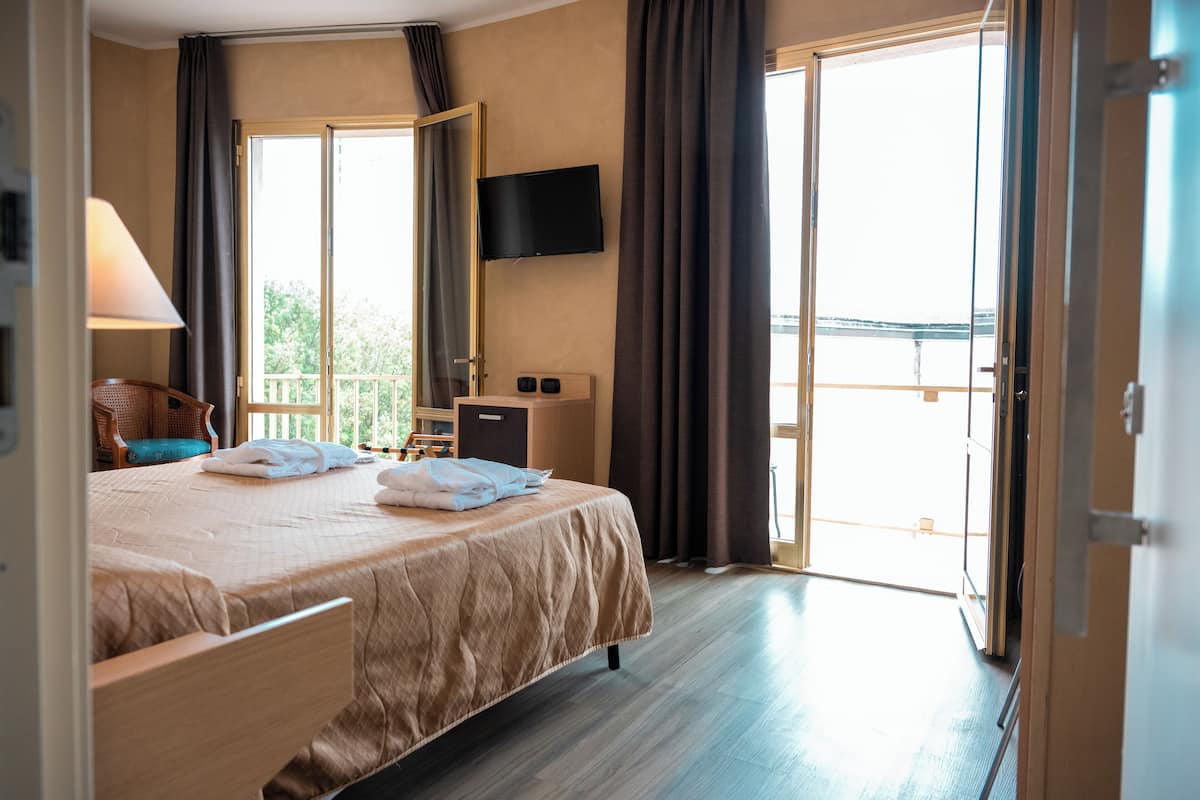 Quarto de hotel com cama, escrivaninha, cadeira, televisão e varanda com cortinas.