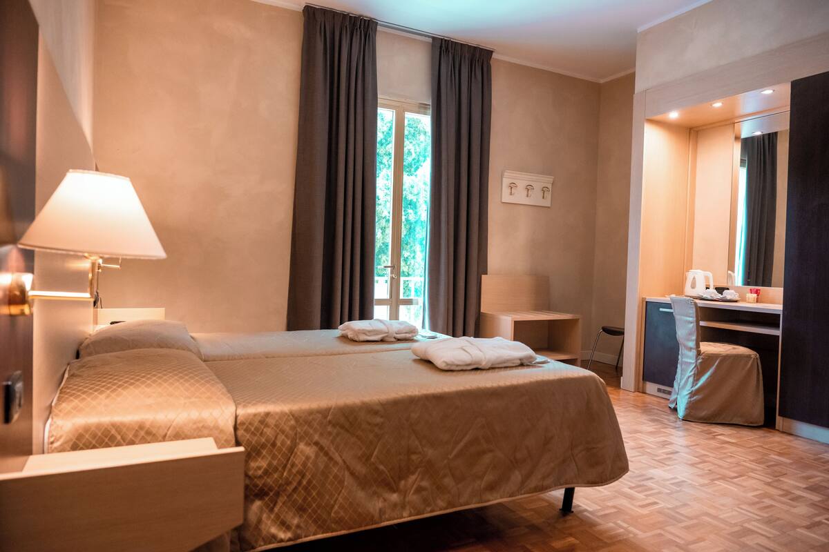 Quarto de hotel com cama grande, escrivaninha, cadeira, luminária e janela com cortinas.