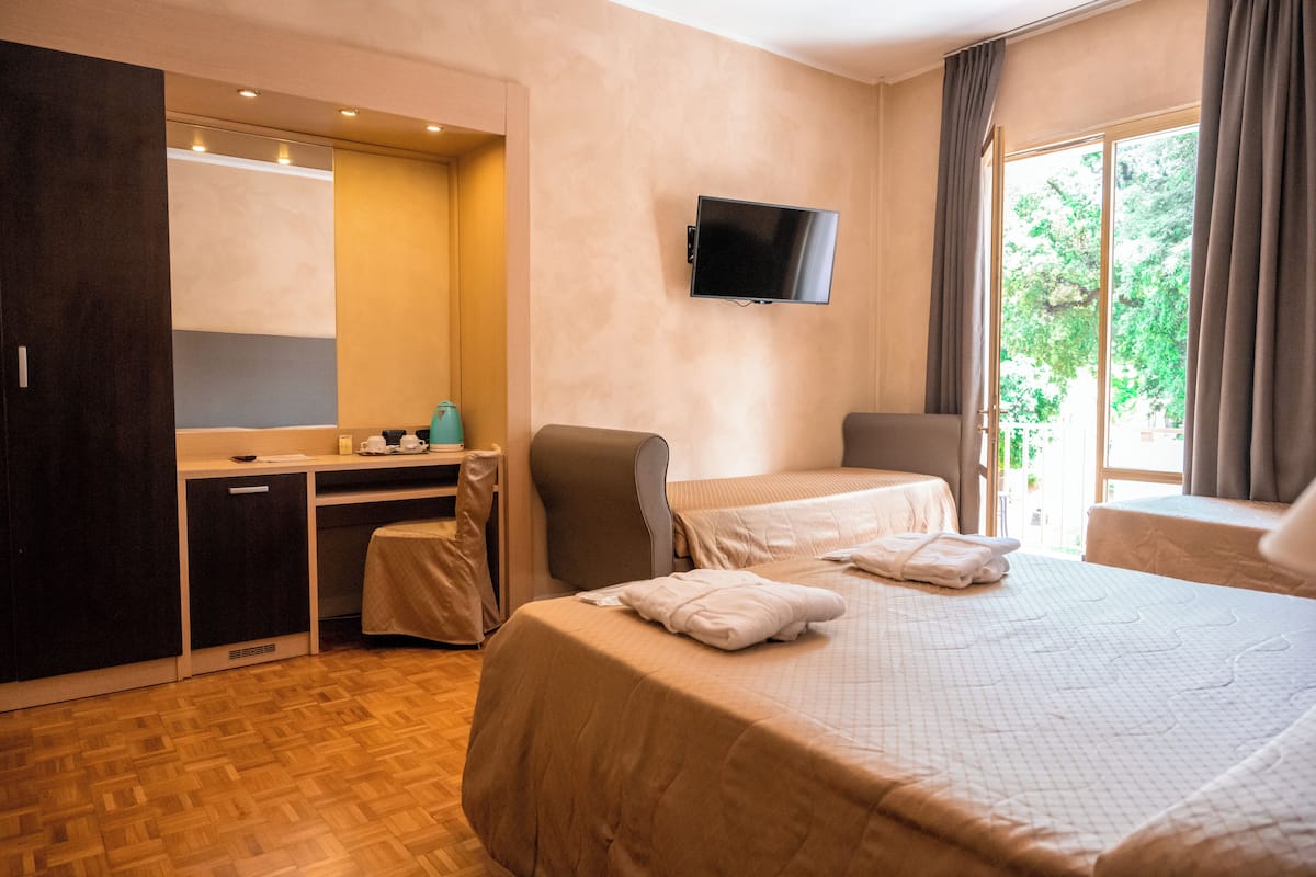 Quarto de hotel com cama, escrivaninha, cadeira, televisão e janela com cortinas.