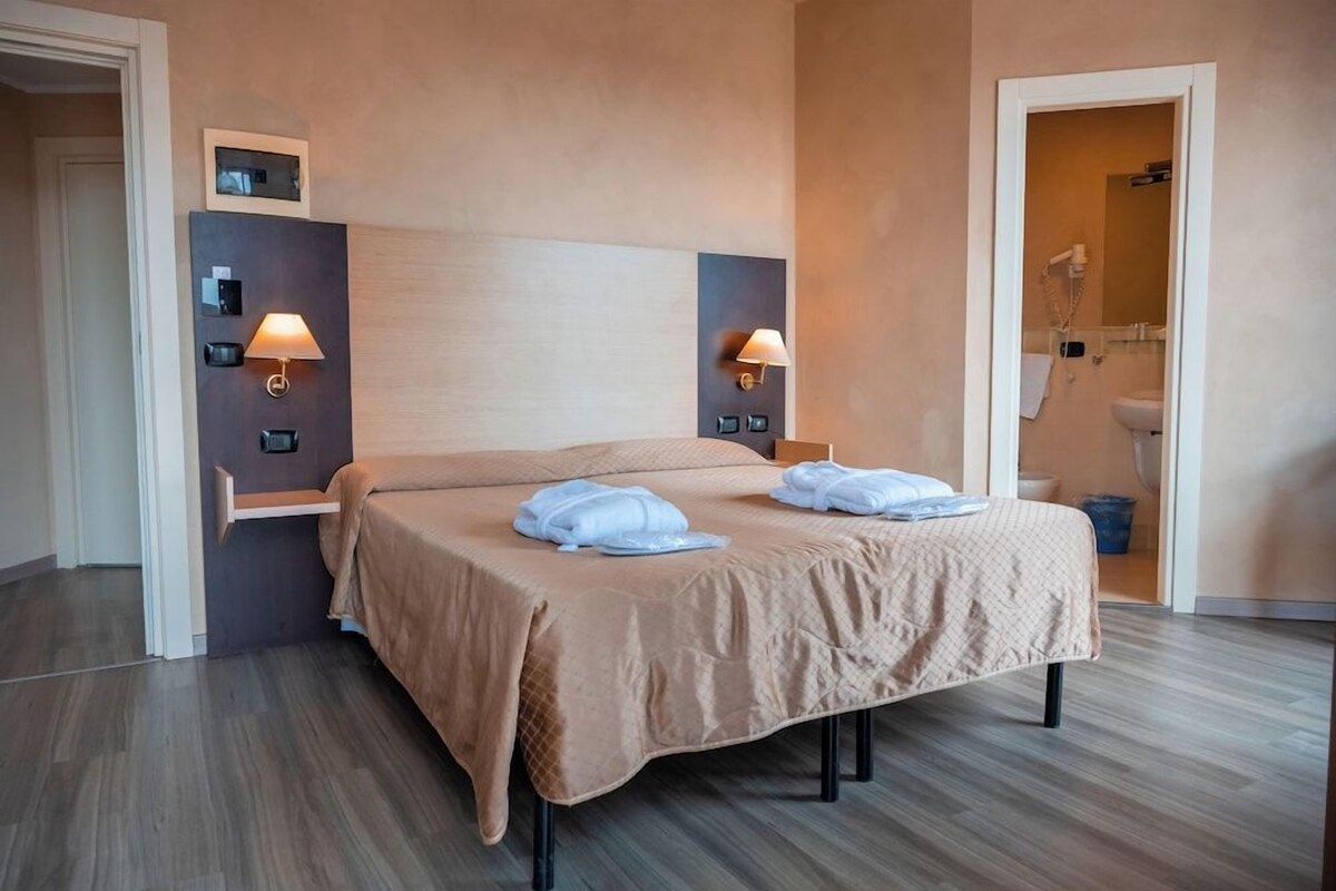 Une chambre d’hôtel avec un lit, des lampes de chevet, un miroir et une salle de bain visible à travers une porte ouverte.