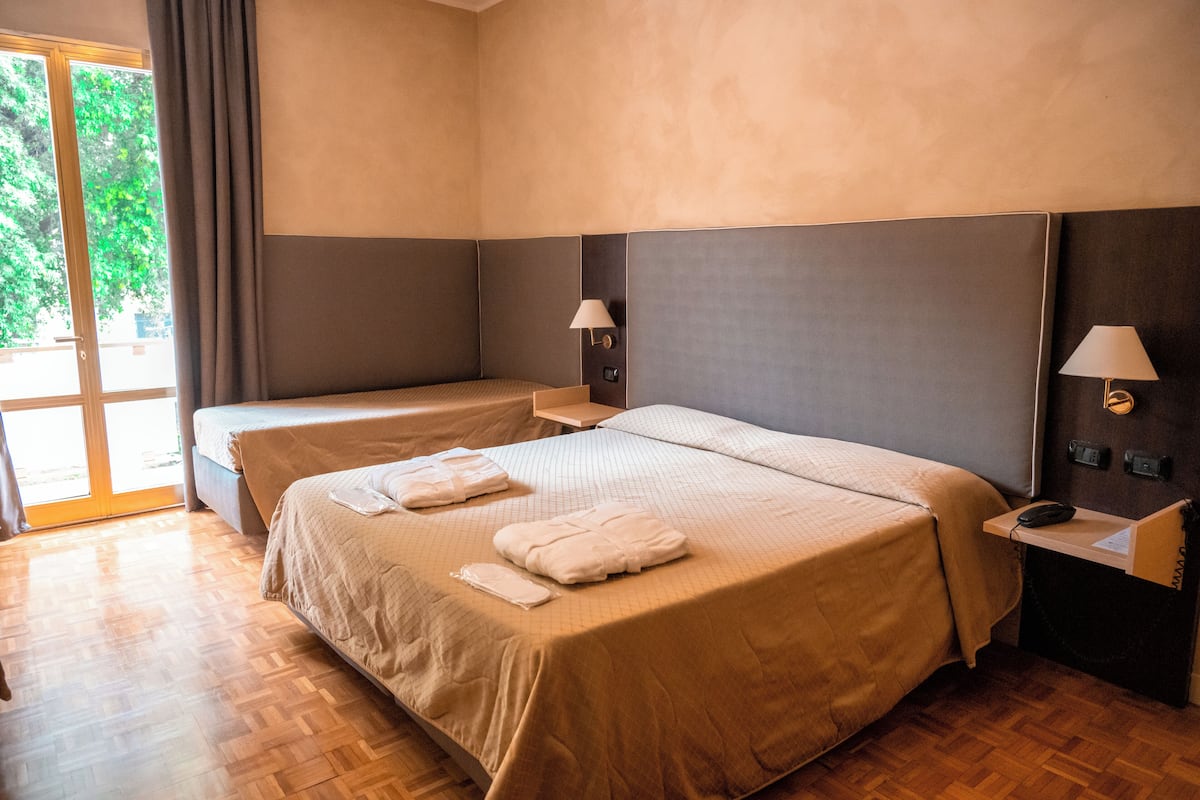 Quarto de hotel com uma cama grande, uma cama menor, uma escrivaninha com abajur e vista para áreas verdes.