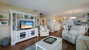 Un soggiorno con televisione, arredamento bianco e una cucina sullo sfondo.