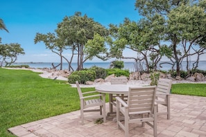 A patio with a table and chairs overlooking a beach with trees and rocks.