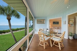 A dining area with a glass table and wicker chairs, overlooking a grassy area with a palm tree and a view of the sea.