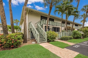 A two-story building with a green staircase leading to the entrance, surrounded by palm trees and well-maintained lawns.