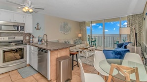 A modern kitchen with a microwave, stove, and dishwasher, a dining area with a glass table and chairs, and a balcony with a view of the sea. - Cozy 2BR/2BA Oceanfront Condo - Steps from the Beach! (Murrells Inlet)