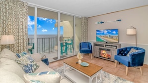 A living room with a sofa, two chairs, a coffee table, a TV, a fireplace, and a balcony with a view of the sea. - Cozy 2BR/2BA Oceanfront Condo - Steps from the Beach! (Murrells Inlet)
