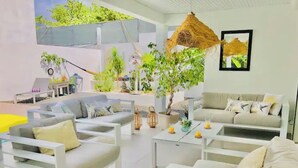 Un espacio interior moderno y bien iluminado con muebles blancos, incluyendo sofás y una mesa de centro, organizados alrededor de una zona central con plantas y una lámpara colgante.