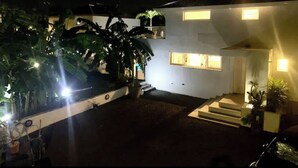 Una casa de dos pisos con fachada blanca, un porche delantero y una pequeña escalera que conduce a la entrada. Hay plantas en macetas y un árbol grande en el jardín delantero.