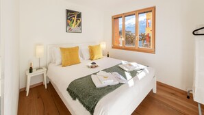 Een net opgemaakt bed met witte lakens, een groene deken en handdoeken gevouwen aan het voeteneinde. Er staat een klein tafeltje met een lamp en een fles champagne. Een ingelijst beeld hangt aan de muur boven het bed.