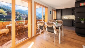 Een moderne keuken met donkere kastjes en een zwarte boekenkast, een zithoek met houten stoelen en een tafel, en een balkon met een houten terras en uitzicht op bergen.