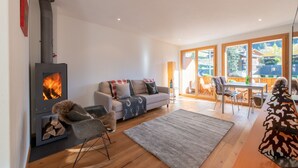 Een moderne woonkamer met een houtkachel, een bank, een eettafel en grote ramen met uitzicht op de natuur.
