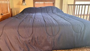 Una cama grande con cubierta azul oscuro en una habitación con puerta de madera, lámpara de pared y ventana con persianas.