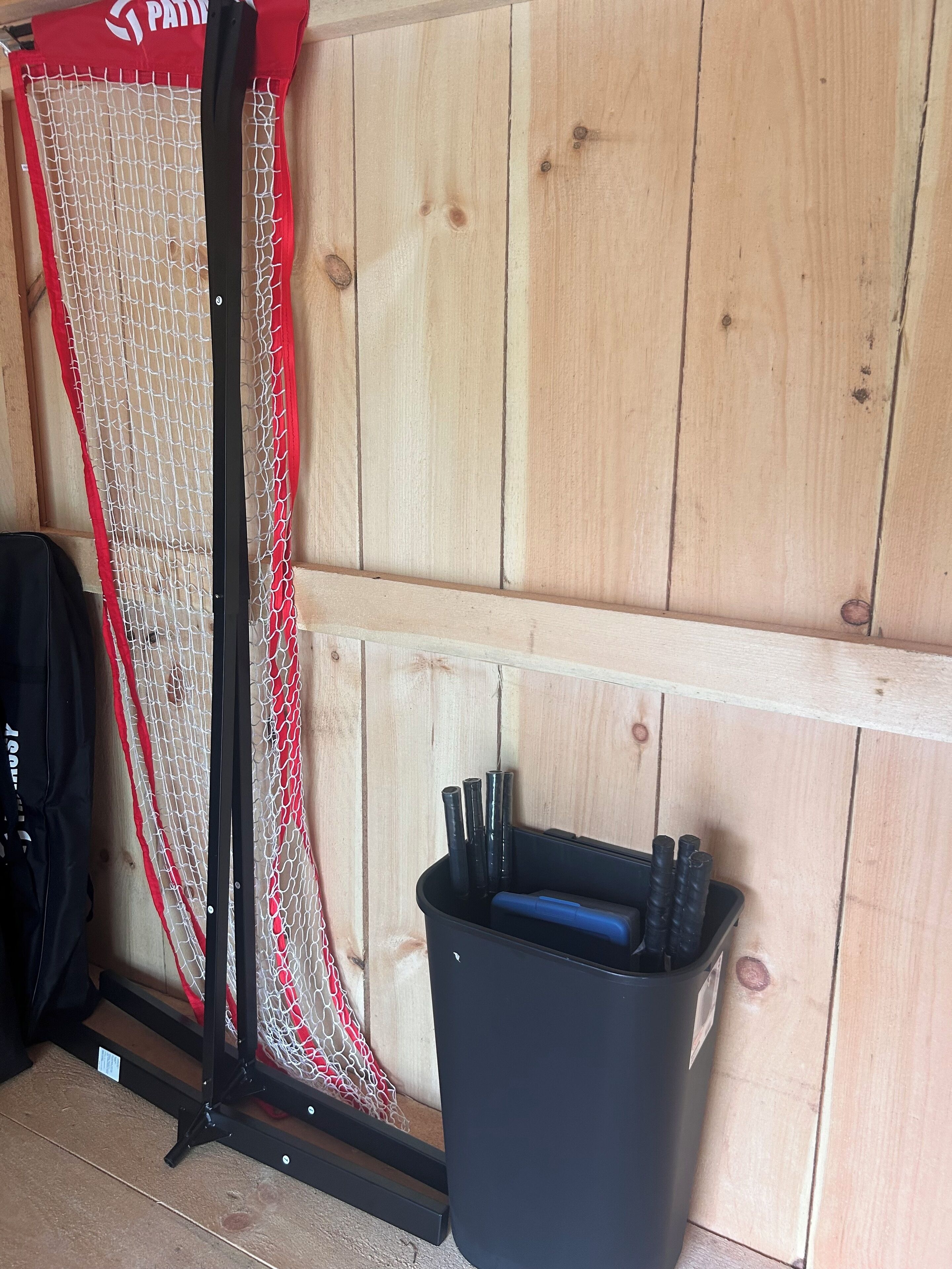 A wooden wall with a red and white netting structure, a black trash bin with tools, and a black bag on the floor.