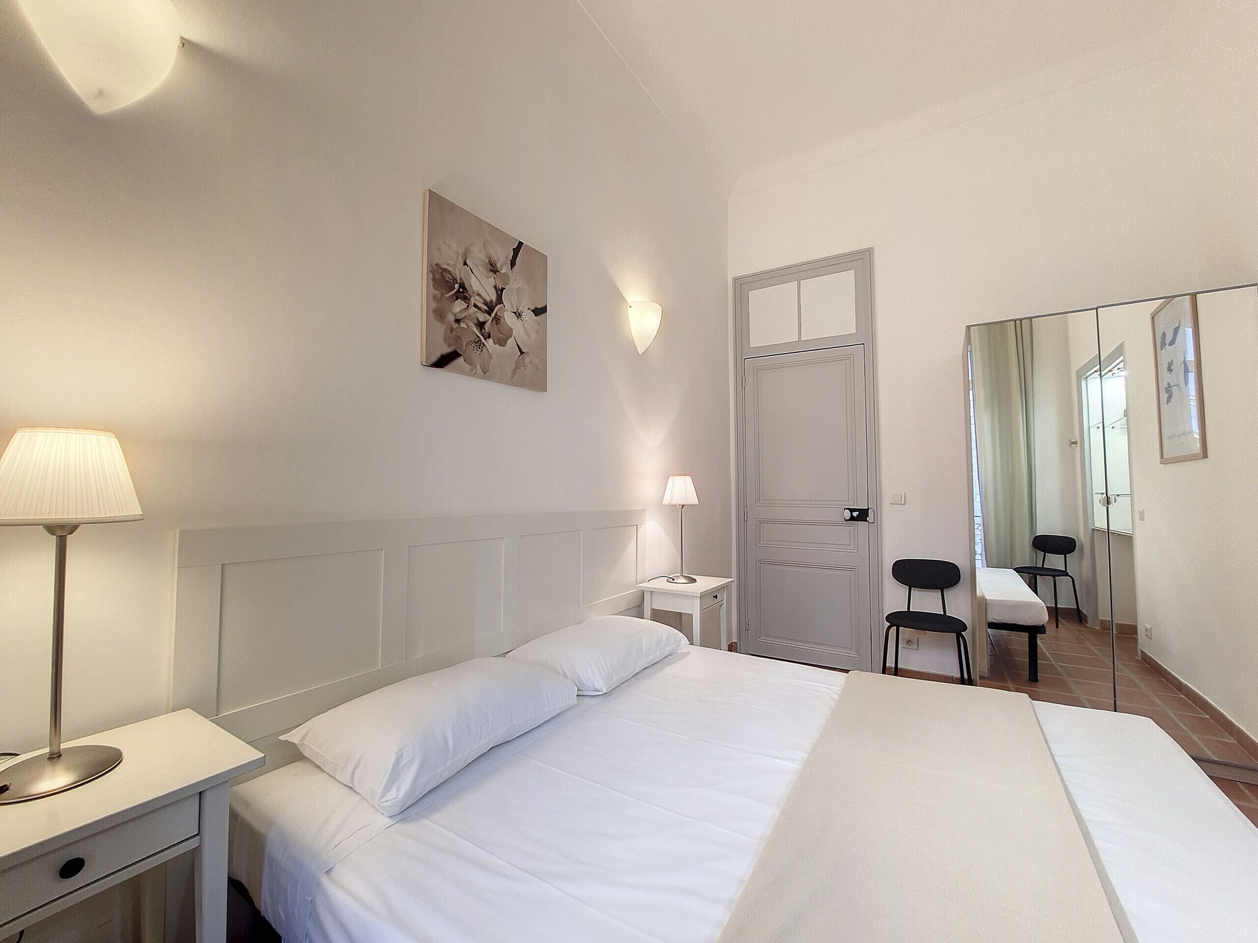 Ein Hotelzimmer mit einem Bett, Nachttischlampen, einem Schreibtisch mit Stuhl, einem Spiegel und einer Tür.