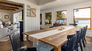A dining area with a wooden table and chairs, white paneled walls, and large windows overlooking a body of water. - Private Sandy Beach, Dock, Kayaks, Canoe & Paddleboat | Sleeps 12 | Dog Friendly (Central Lake)