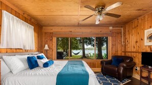 A wooden cabin room with a bed, a brown leather chair, a TV, and a large window offering a view of the outdoors. - Private Sandy Beach, Dock, Kayaks, Canoe & Paddleboat | Sleeps 12 | Dog Friendly (Central Lake)