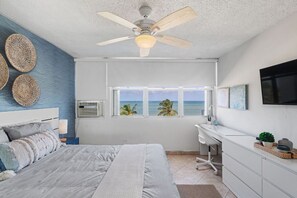 Een slaapkamer met een bed, een bureau, een plafondventilator, een raam met uitzicht op de oceaan en een televisie aan de muur.