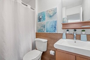 Een badkamer met douchegordijn, toilet, wastafel met spiegel en een muur met een blauw abstract schilderij.