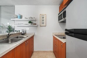 A modern kitchen with wooden cabinets, a stainless steel sink, a black refrigerator, and a microwave. There is a small potted plant on the countertop and a framed picture on the wall.