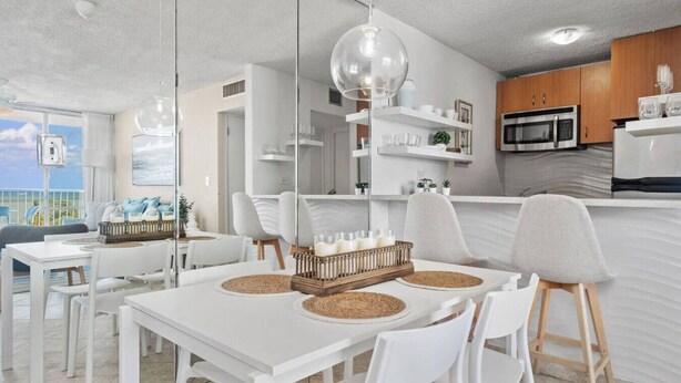 Una cucina moderna con mobili bianchi, un forno a microonde e un frigorifero. C'è una zona pranzo con un tavolo e sedie bianche, e una grande finestra con vista sull'oceano.
