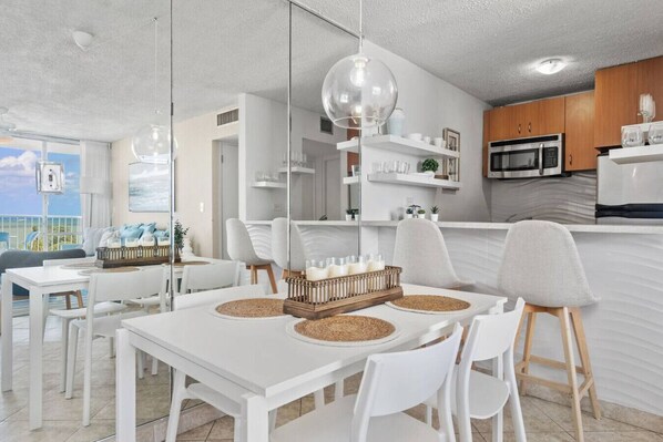 A modern kitchen with white cabinets, a microwave, and a refrigerator. There is a dining area with a white table and chairs, and a large window offering a view of the ocean.