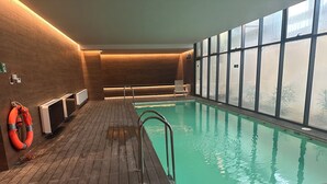 Piscina interior com piso de madeira, um flutuador de segurança e vista através de grandes janelas.