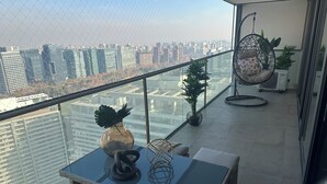 Varanda moderna com guarda-corrimão de vidro, cadeira suspensa, mesinha com objetos decorativos e plantas em vasos.