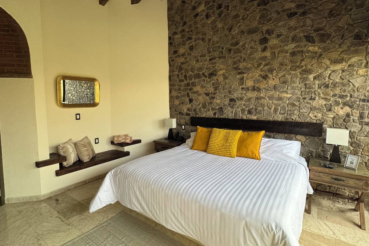 Un dormitorio con pared de piedra, una cama con ropa de cama a rayas, cabecero de madera y un pequeño estante con almohadas.