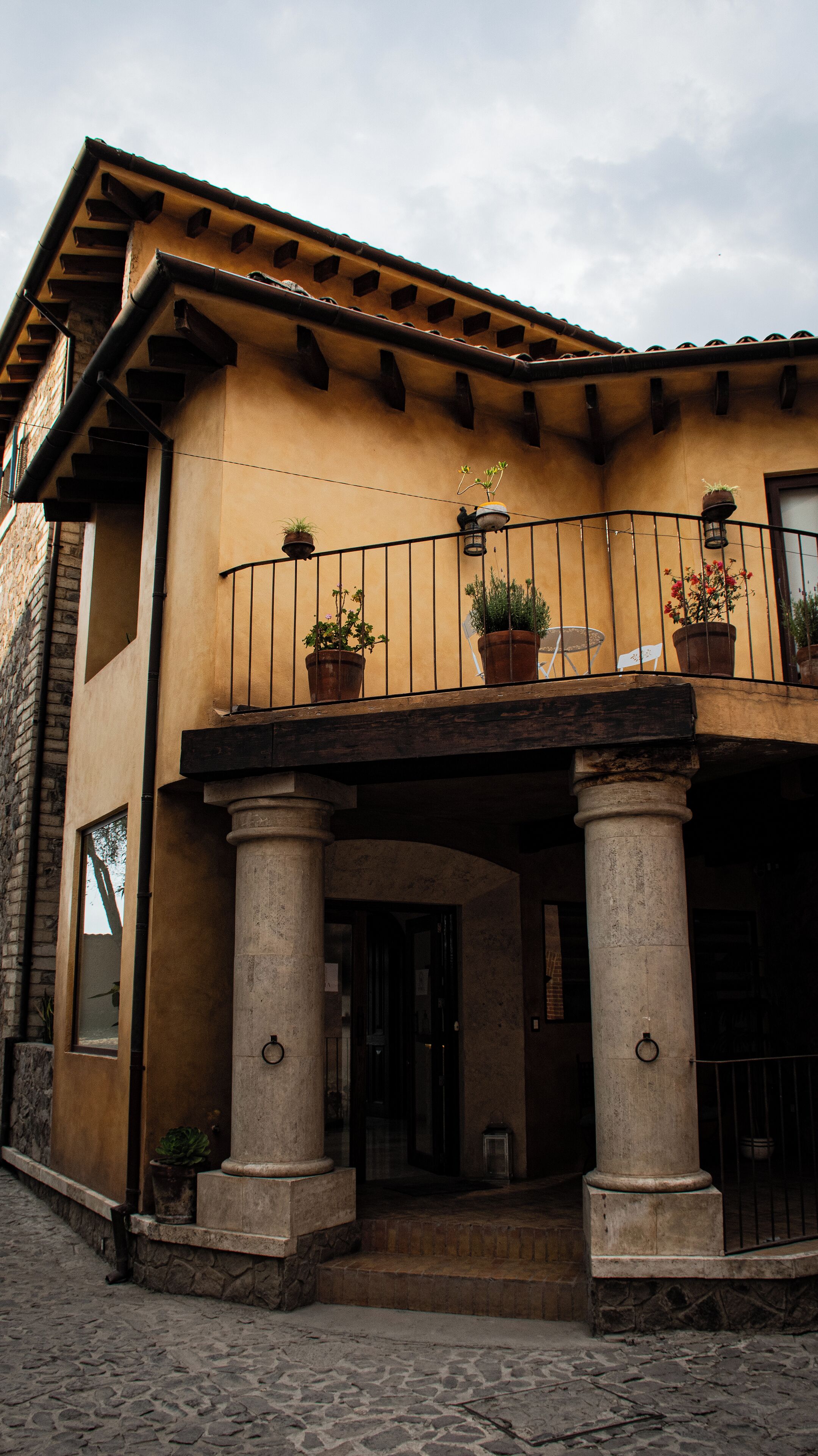 A building with a stone facade, a balcony with potted plants, and a cobblestone entrance.