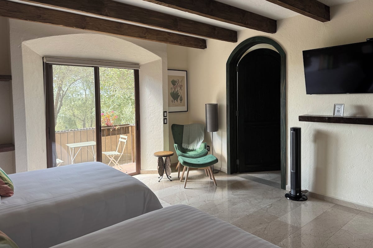 Habitación de hotel con dos camas, un balcón con mesa y sillas, una silla verde, un televisor y una puerta.