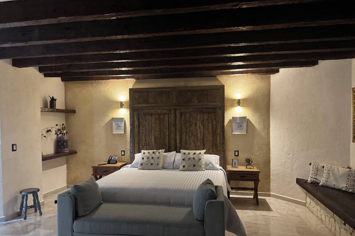 Un dormitorio con cama, sofá, vigas de madera, estantes con objetos decorativos e iluminación empotrada.