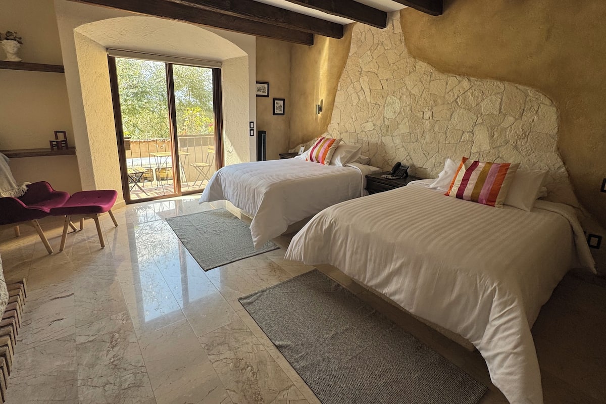 Habitación de hotel con dos camas, un balcón, una silla y una pared de piedra.