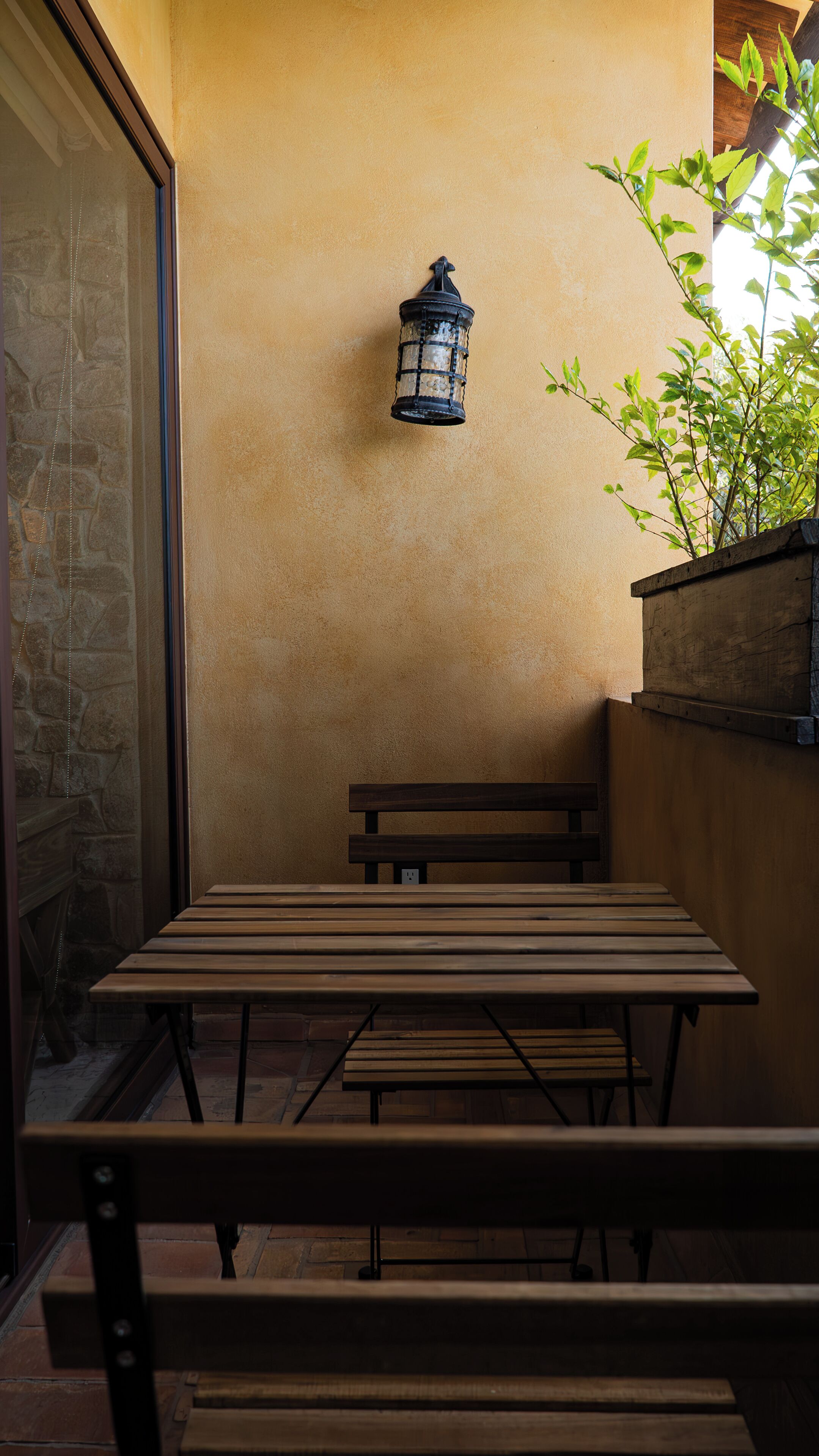 Un banco y una mesa de madera frente a una pared con textura, con un farol y una maceta con follaje verde.