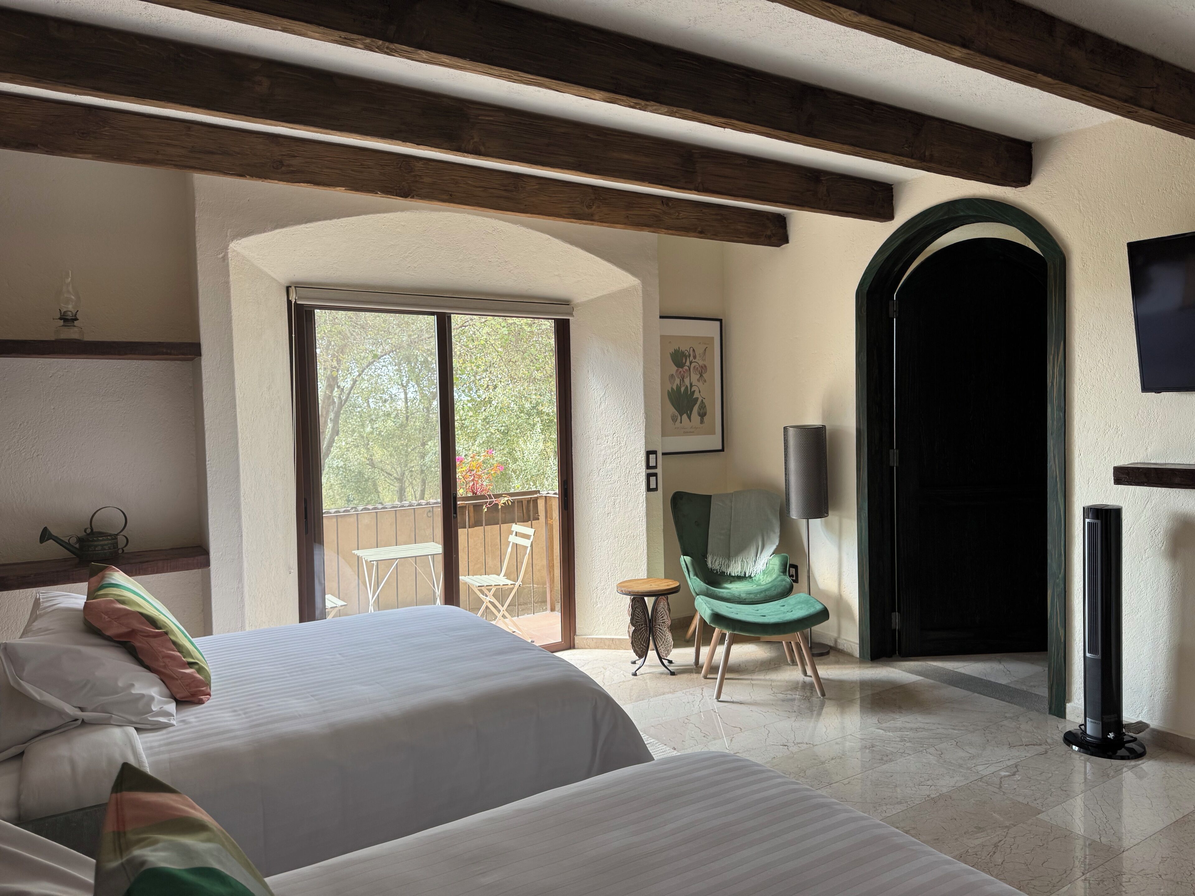 A hotel room with two beds, a green armchair, a small table, a floor lamp, and a large arched doorway leading to a balcony.