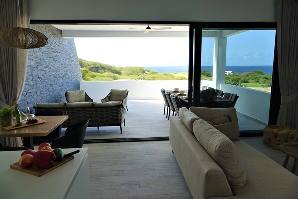 Ein modernes Wohnzimmer mit Sofa, Esstisch und Stühlen, ein Balkon mit Meerblick und ein großes Fenster.