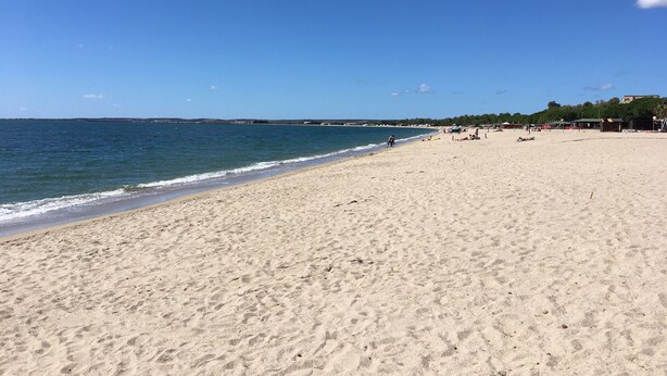 Ein weiter Sandstrand mit sanften Wellen, blauem Himmel und Gebäuden in der Ferne.