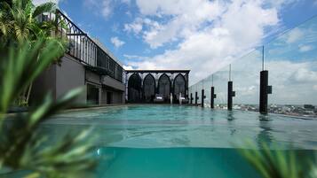 Una piscina infinita en la azotea con vistas a un horizonte urbano, un edificio moderno con ventanas arqueadas y exuberante vegetación.