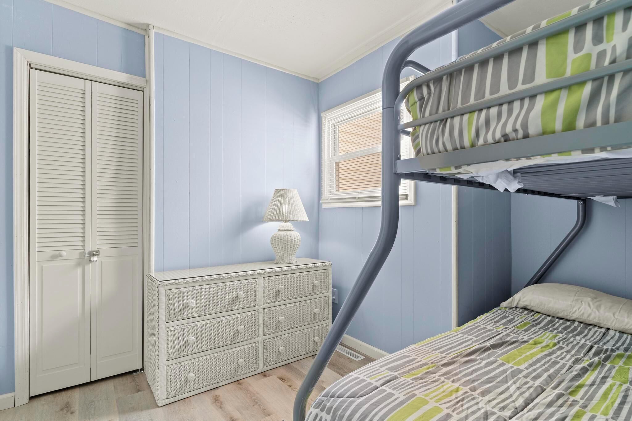 A bunk bed with a striped bedspread, a white dresser with drawers, a lamp, and a closed door.