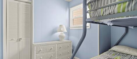 A bunk bed with a striped bedspread, a white dresser with drawers, a lamp, and a closed door.