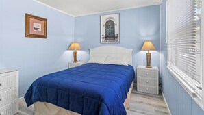 Un dormitorio con colcha azul, dos lámparas de noche, un cuadro enmarcado y una ventana con persianas.