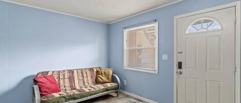 Una habitación con pared azul claro, una puerta blanca, una ventana con persianas y un sofá con cojines estampados.