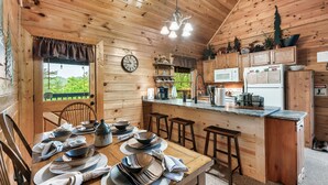 Ein Holzblockhaus-Innenraum mit Esstisch, gedeckt für vier Personen, einer Küchenzeile mit Kühlschrank, Mikrowelle und Kaffeemaschine sowie einem großen Fenster mit Blick auf Grünflächen.