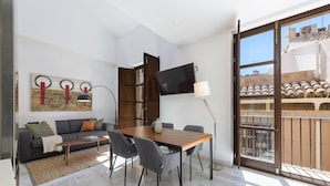 Una sala de estar moderna con un sofá, una mesa de centro y una mesa de comedor. Hay un televisor montado en la pared y una ventana con vistas a edificios.