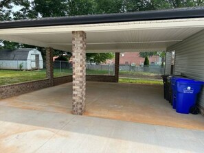 Ein überdachter Patio-Bereich mit einer Ziegelpfeiler, einem Betonboden und einer Reihe blauer Recyclingbehälter.
