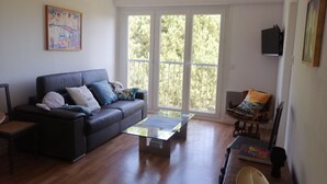 A living room with a dark leather sofa, a glass coffee table, and a wooden side table. There is a large window with a view of trees and a door to the right. - Appartement Plage et Lac, 4pieces 3 Chambres (Les Sables-d'Olonne)