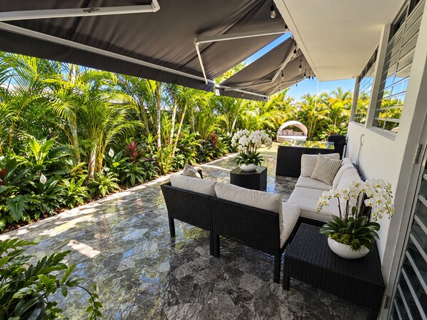 An outdoor patio with a sofa and table, surrounded by lush tropical plants and a clear view of the sky. - Beach House with Private Pool, 15 minutes from the airport. Tourist area. Places. (Loíza)