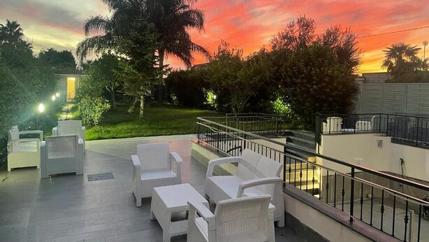 Un área de estar al aire libre con mobiliario de mimbre blanco, un balcón con barandilla y vista al atardecer.