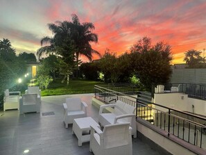 Un espace extérieur aménagé avec des meubles en osier blanc, un balcon doté d’une balustrade et une vue sur le coucher de soleil.